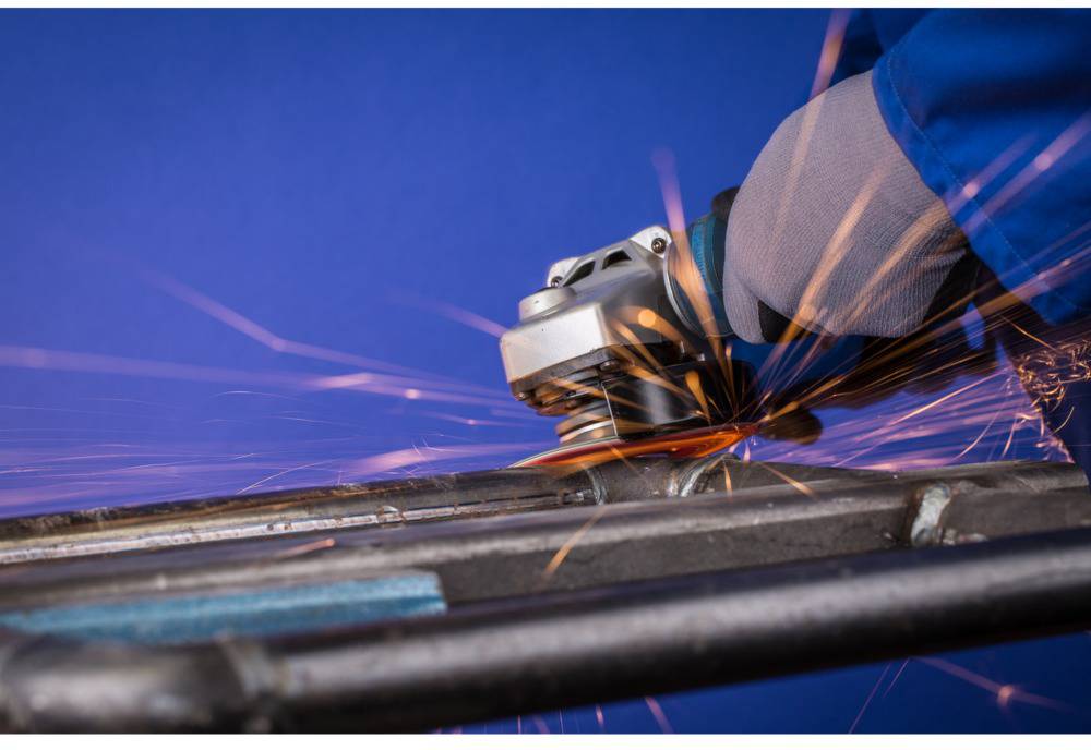 A person is grinding metal with an angle grinder, sparks flying. The background is blue. Protective clothing is being worn.