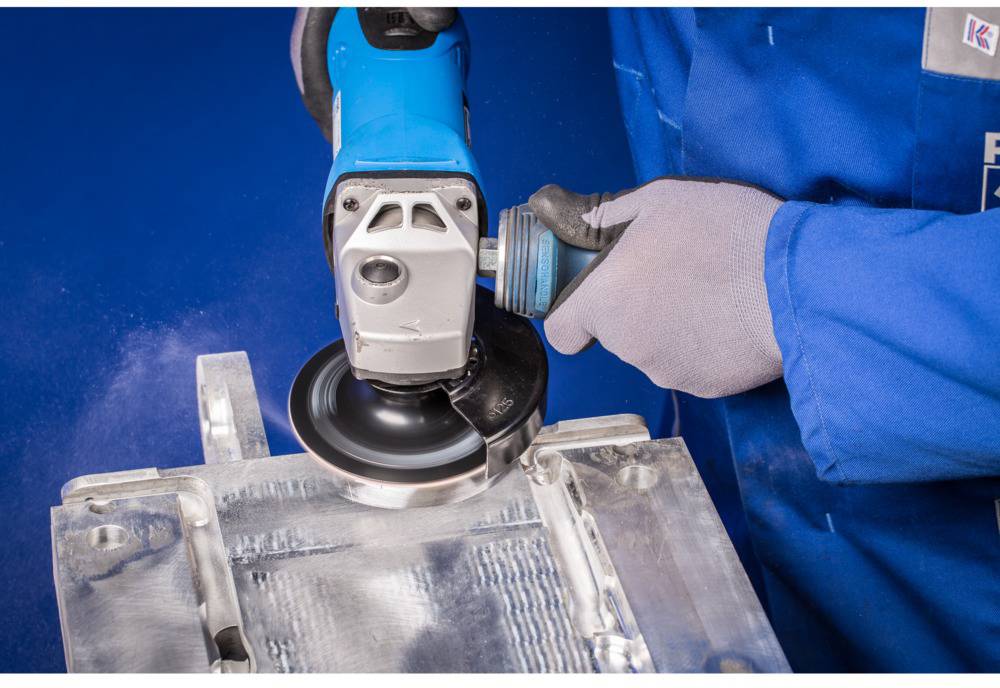 A person in blue workwear is grinding a metal surface with an electric grinding machine. Protective clothing is being worn.