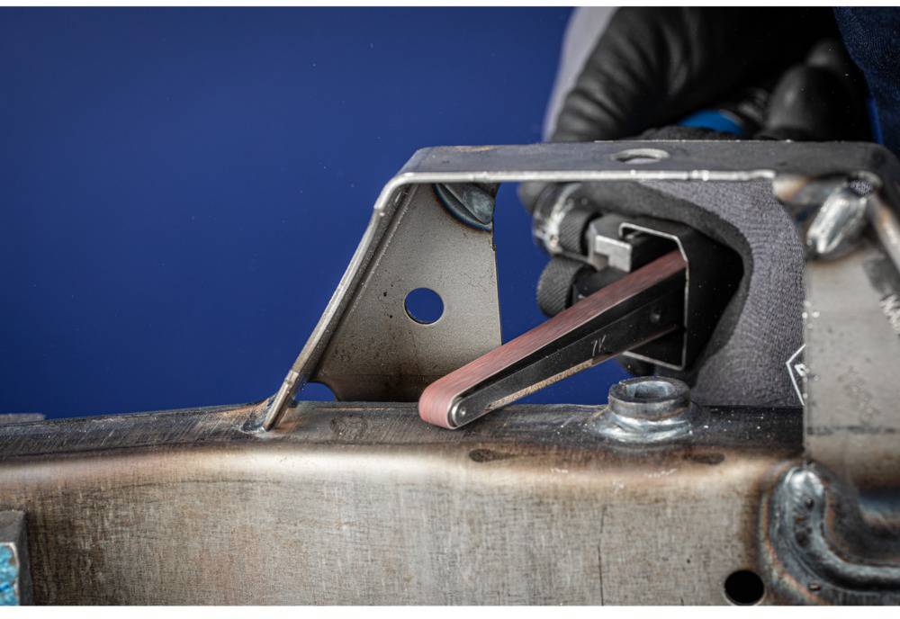 Close-up of a metal grinding machine working on a steel workpiece. A gloved hand guides the tool.