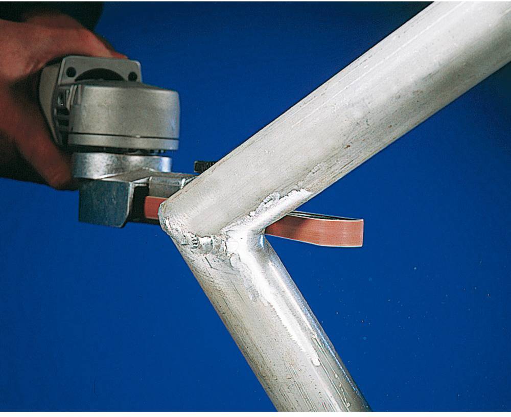 A grinding machine grinds a metal edge with an abrasive belt. Hands are holding the device, working on the corner of a metal pipe.