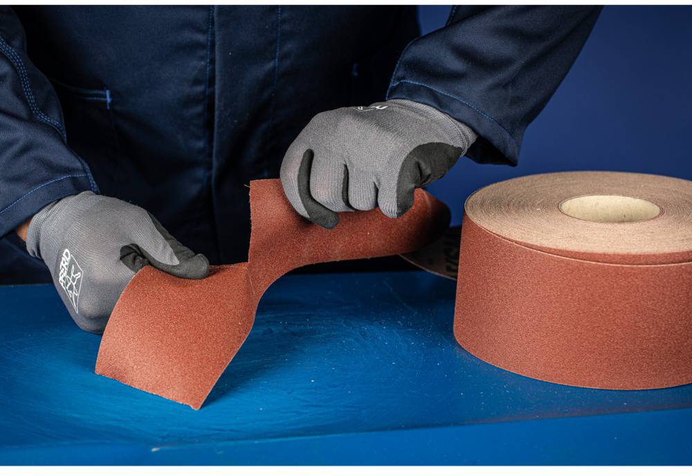 A person wearing grey gloves is cutting red sandpaper from a roll.