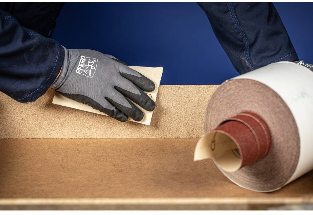 A person wearing gloves is sanding a wooden surface with sandpaper. Beside them is a roll of sandpaper.