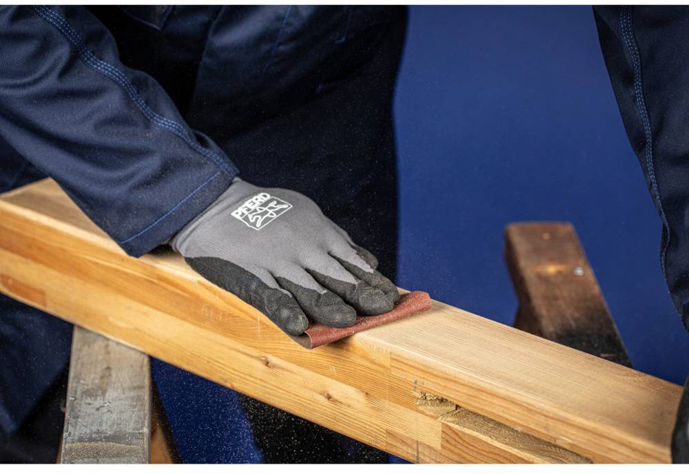 A person is sanding a wooden board with sand-coloured sandpaper. They are wearing grey gloves and dark workwear.