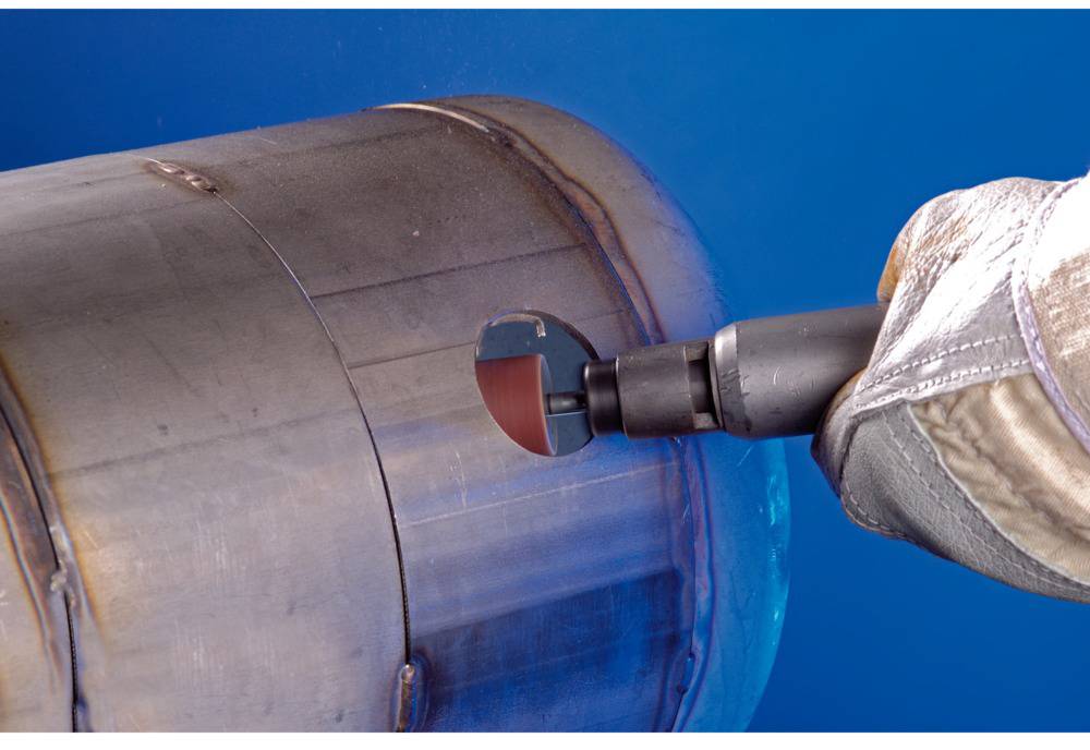 A person is grinding the outer surface of a metal cylinder with a grinding tool. Protective clothing is being worn.