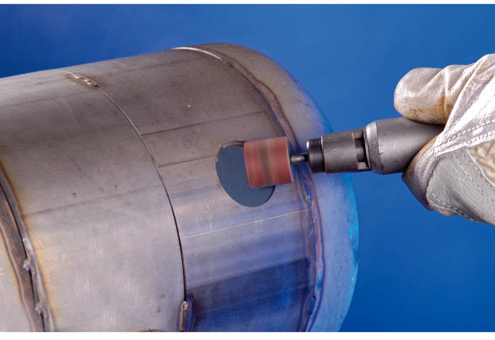 A craftsman is grinding rust off a metal cylinder with an angle grinder to smooth the surface. Hands wearing safety gloves.