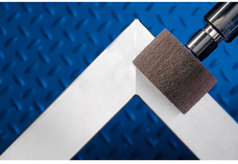 A metal sheet is polished using a grinding tool. The background shows a blue, diamond-shaped pattern.