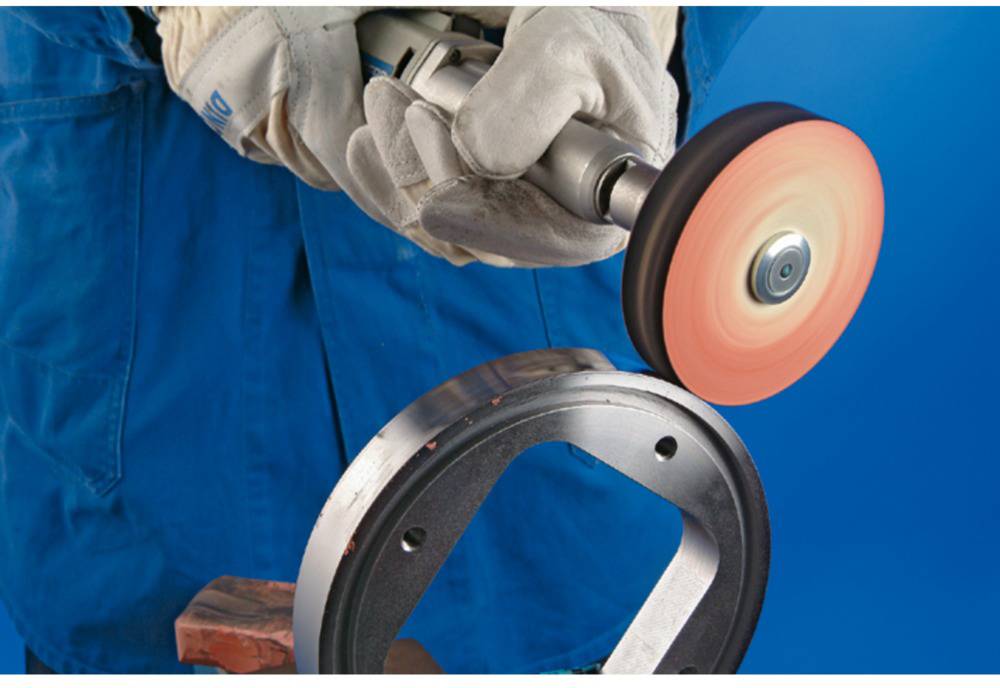 A person in blue workwear is grinding a circular metal piece with a rotating disc. Wearing gloves, focused on precision.