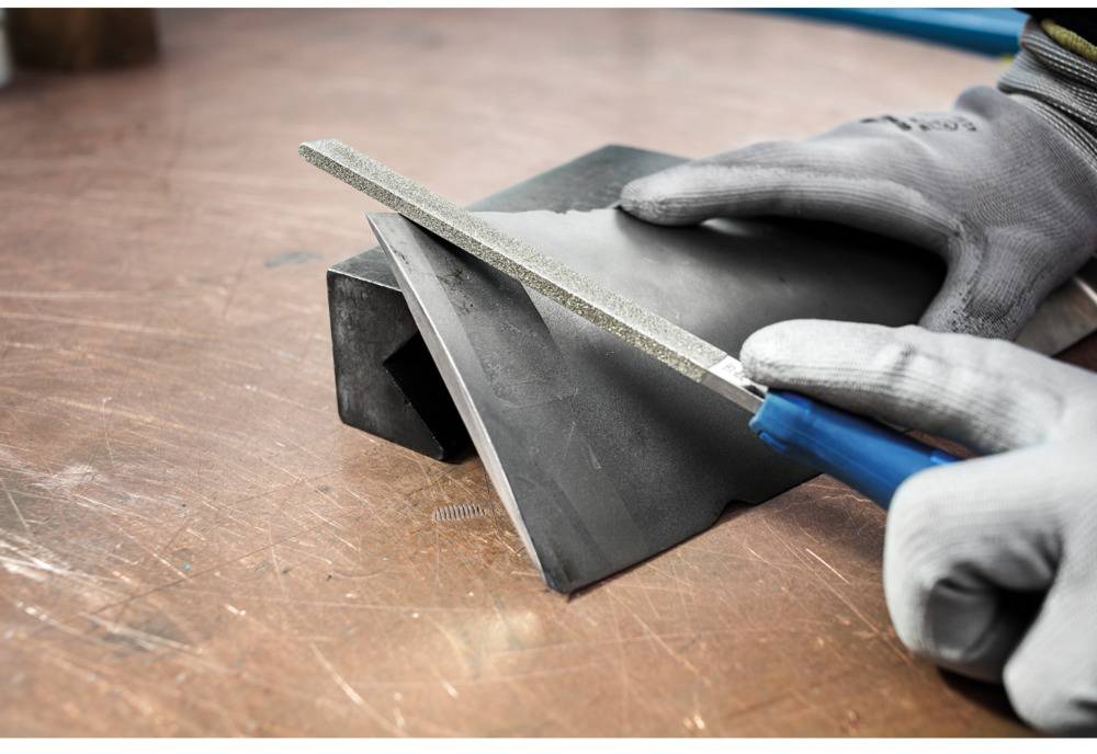 A person is filing a piece of metal with a blue file. The hands are protected by grey gloves.