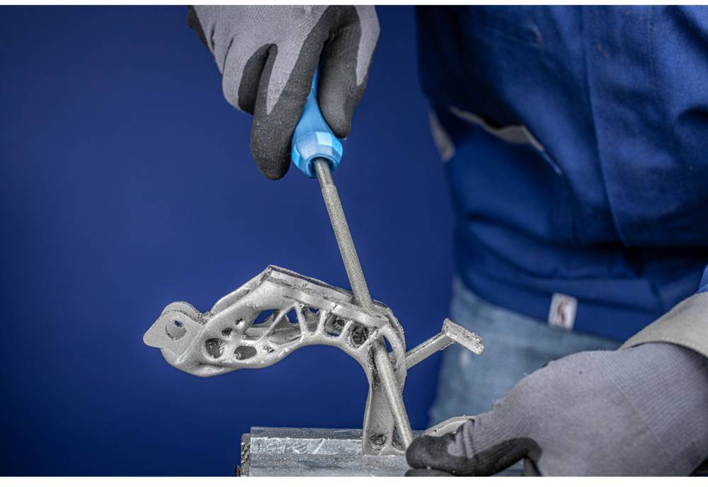 A person is filing a complex metal piece that is clamped in a vice. The person is wearing gloves and a blue jacket.