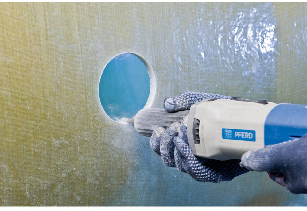 A person is using an electric tool to drill a hole in a yellow, fibreglass-like surface. The tool is blue.