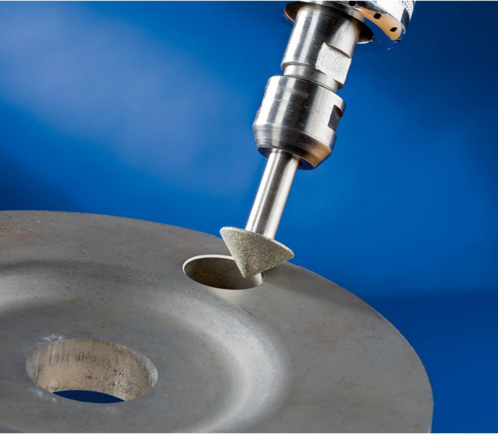 A metallic grinding cone is machining a round opening in a metal plate against a blue background.