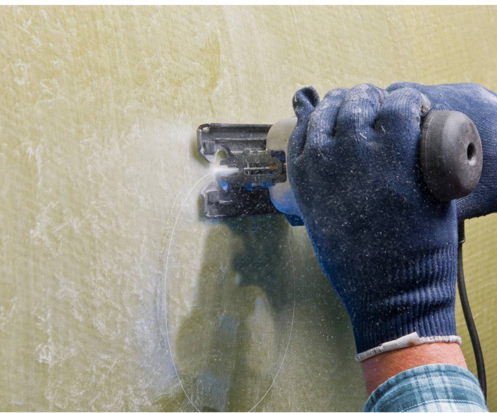 A person is cutting an oval hole in a plasterboard wall using an electric saw. They are wearing blue-grey work gloves.