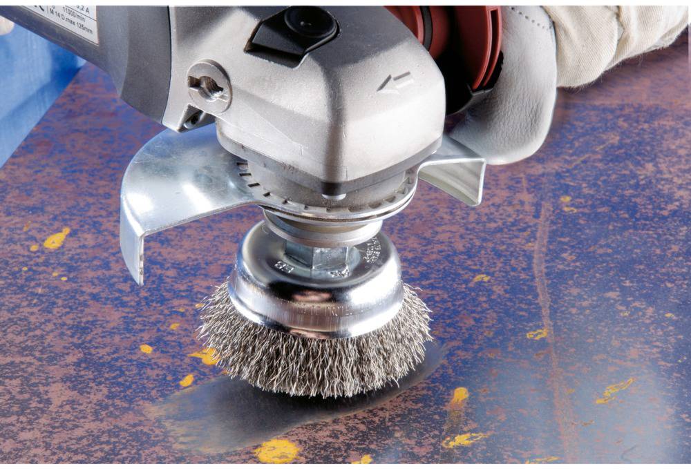 A hand is holding an electric wire brush, removing rust from a metal surface. The surface is rust-brown with blue areas.