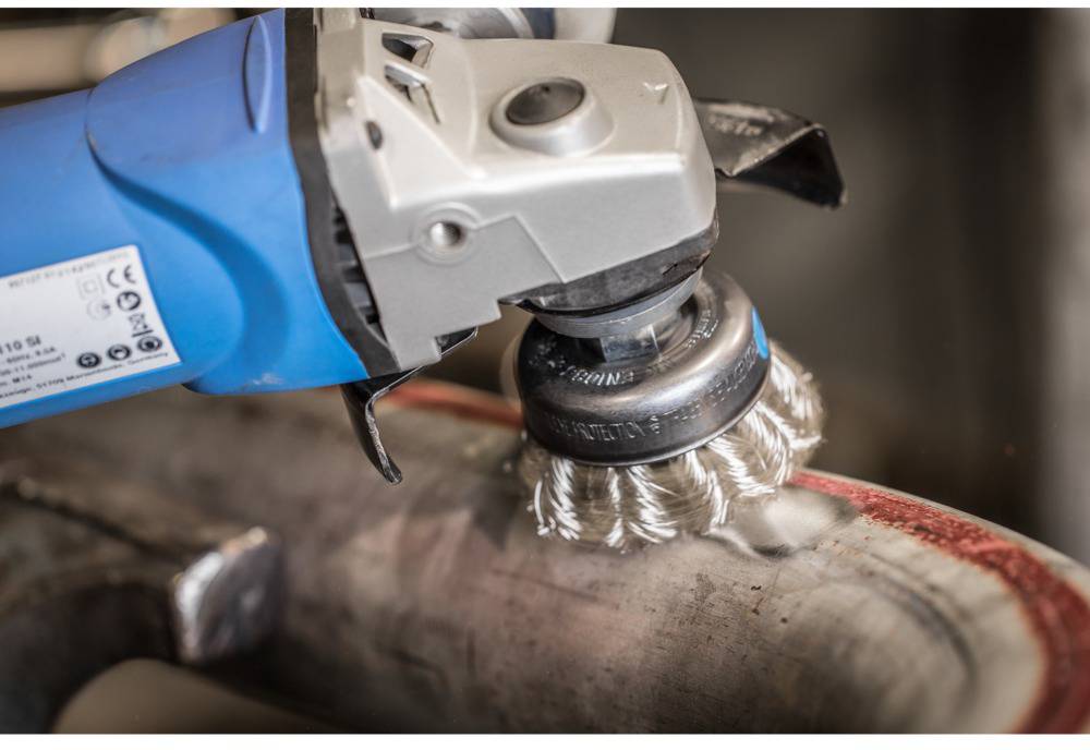 A angle grinder with a wire brush attachment is cleaning a metal surface. The grinder is blue and visible in action.