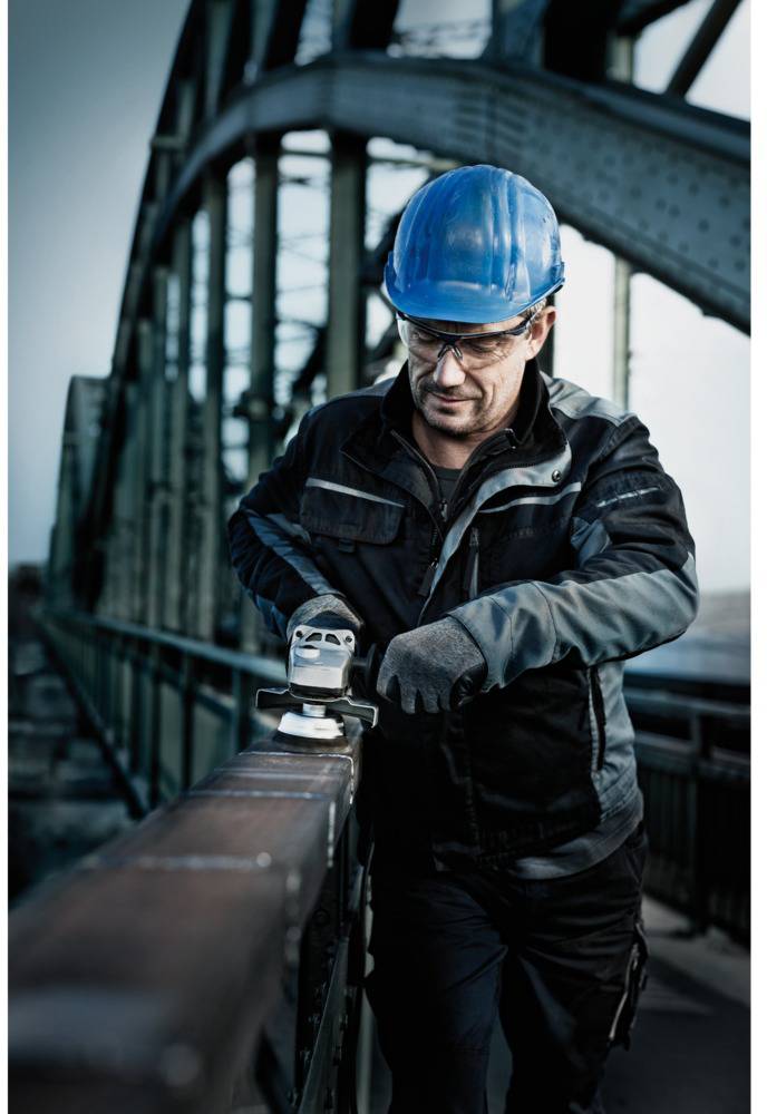 A man wearing safety glasses and a hard hat is working with a grinding tool on a metal handrail on a bridge.