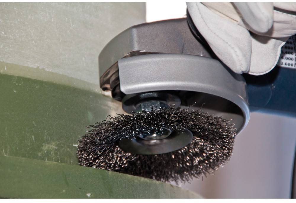 A wire brush on an angle grinder cleans the surface of a green pipe. A glove holds the tool securely.