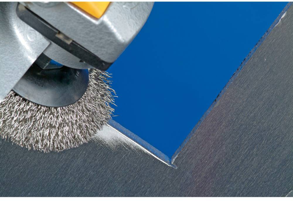 Grinding machine with wire brush processing the corner of a metal plate, blue surface in the background.