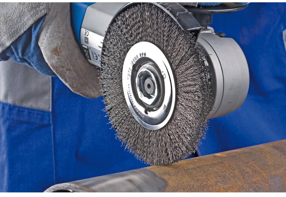 A person is grinding rust off a metal pipe with an electric wire brush. The hand is wearing a work glove.