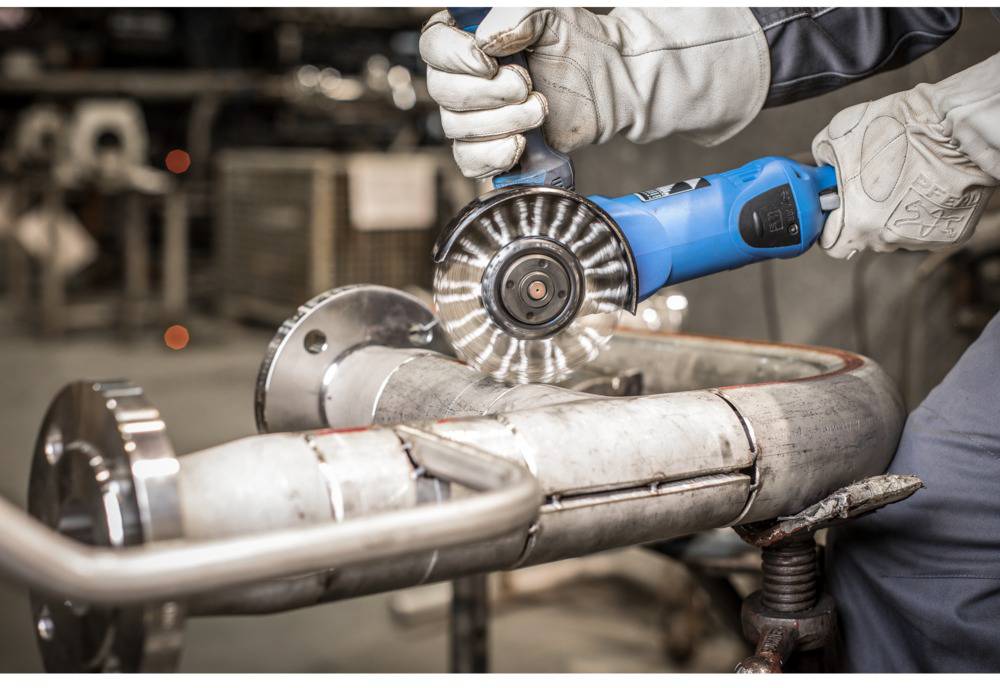 A person is polishing a metal pipe with a blue angle grinder. Safety equipment such as gloves is being worn.
