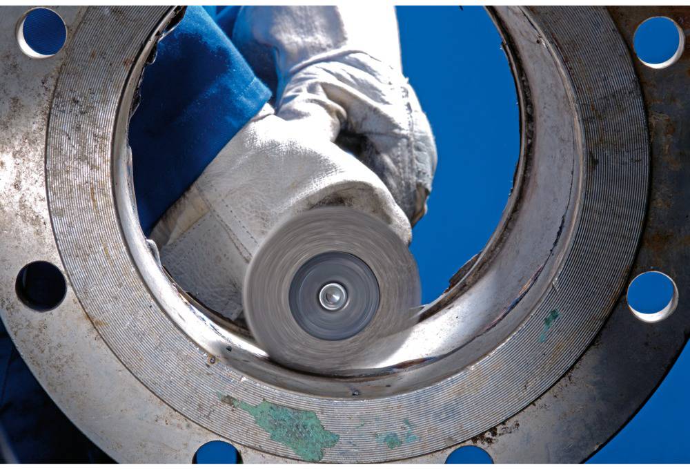 'Worker grinding metal pipe with angle grinder, close-up. The worker is wearing a blue jacket and protective gloves.'
