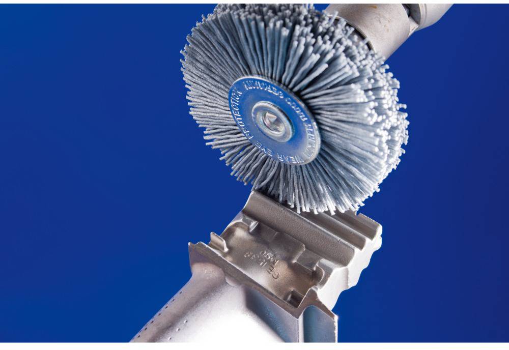 A wire brush is working on a metal surface. The brush is rotating and is close to the edge of the metal. The background is blue.
