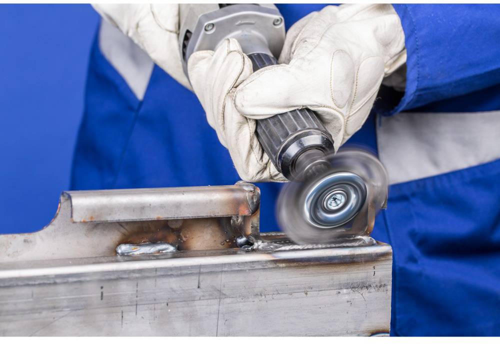 A person wearing protective gloves and blue workwear is grinding a metal edge with an electric grinding machine.