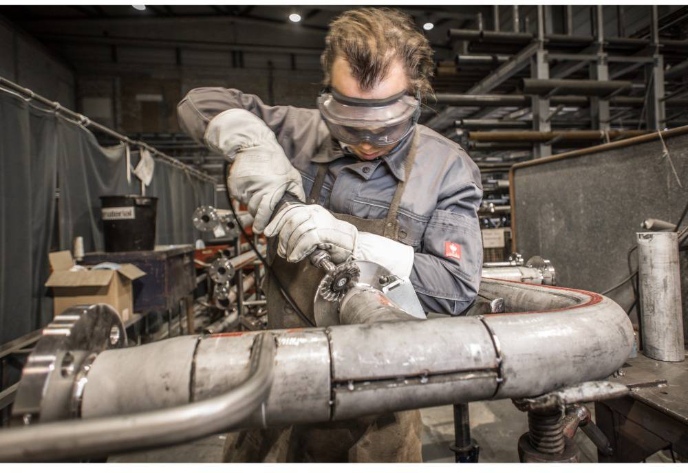 A tradesman is wearing safety glasses and gloves while working on a pipe in a workshop. Tools and materials are scattered in the background.