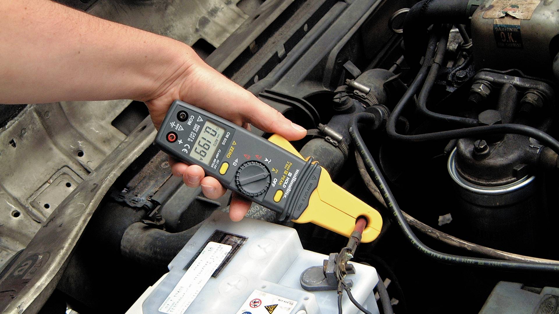 A hand is holding a multimeter set to 12.6 volts, measuring the voltage of a car battery in the engine compartment.