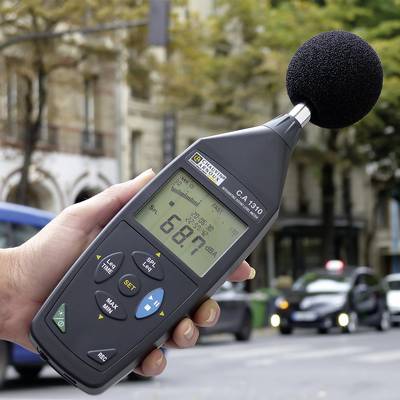 A hand is holding a sound level meter displaying 68.7 dB, on a city street with cars passing by and trees in the background.