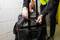 A person pulls the handle of a black suitcase in a storeroom with a fire extinguisher on the wall in the background.