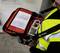 A person wearing a yellow high-visibility vest is holding an open first-aid kit equipped with cables and a digital device.