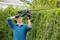 A person wearing safety glasses and gloves is trimming a green hedge with an electric hedge trimmer in the garden.