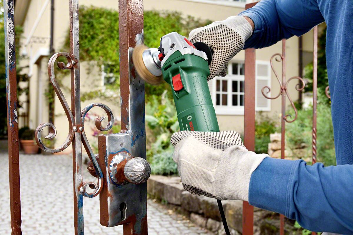 A person is grinding rust off a metal garden gate using a green grinding machine, wearing gloves and blue clothing.