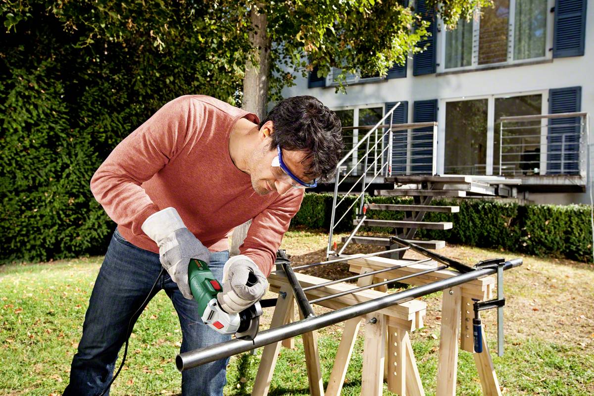A man in a red jumper and safety glasses is cutting a metal pipe outdoors with an electric tool. A modern building is visible in the background.