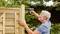 An older man is installing a wooden panel with a cordless drill in the garden, surrounded by green trees and shrubs.