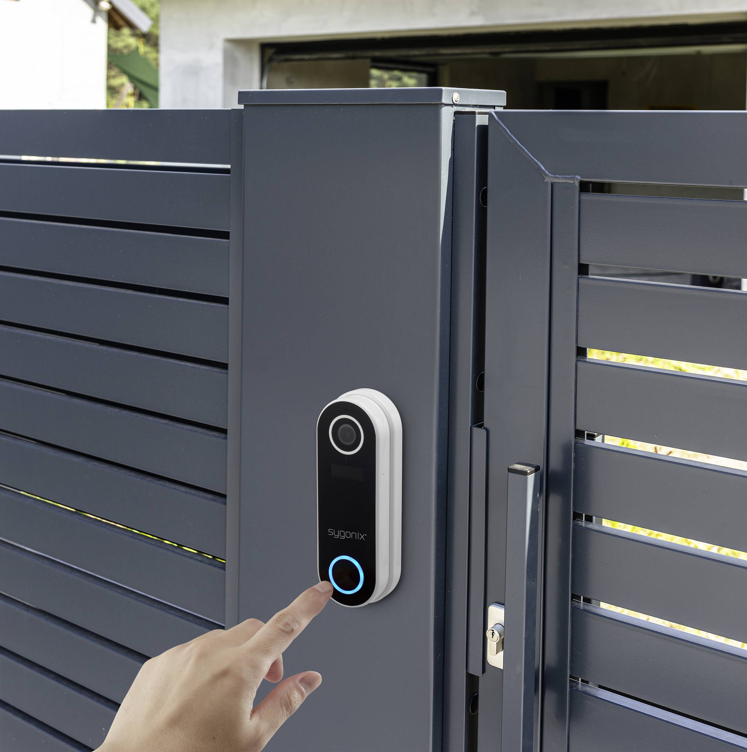 A hand presses the doorbell on a modern, grey garden gate. A building is situated in the background.