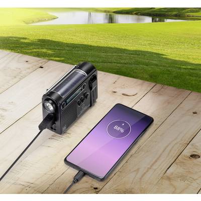 A smartphone is being charged on a wooden table with an outdoor solar charger. A green landscape is visible in the background.