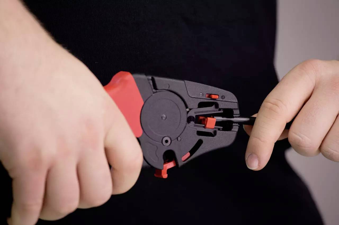 A person is holding a black and red tool, which is being used to work on a black cable. The tool is being operated with both hands.