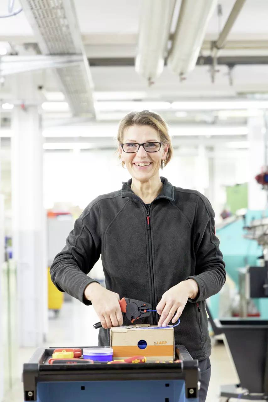 A person in a workshop, smiling at the camera, holding tools, standing at a workbench with a cardboard box and spanner, in a bright environment.