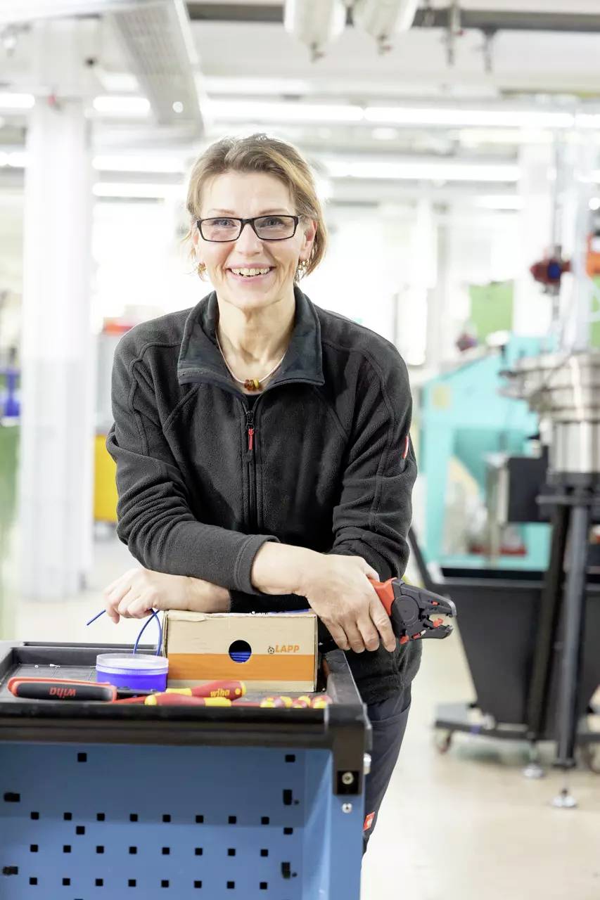 A woman in workwear is smiling and standing in front of a workshop trolley with tools in a workshop environment.