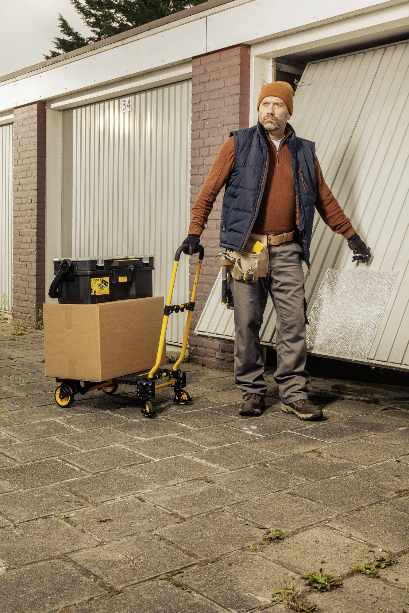 A man opens a garage door and holds a platform with a cardboard box and toolbox. He is wearing work clothes and a cap.