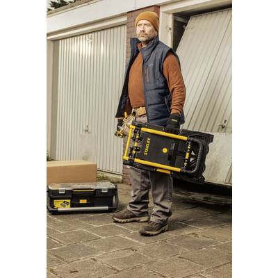 A man in workwear stands in front of garage doors, holding a yellow toolbox and a black box.