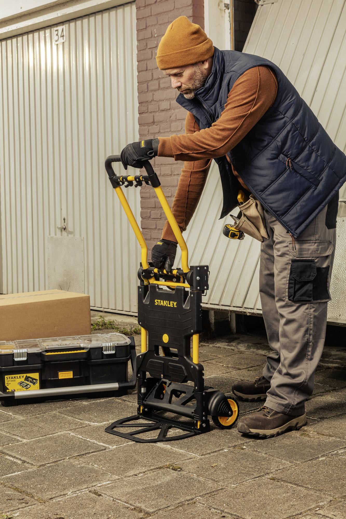 A man wearing a blue waistcoat and orange cap is using a black trolley to transport a crate in a garage driveway.
