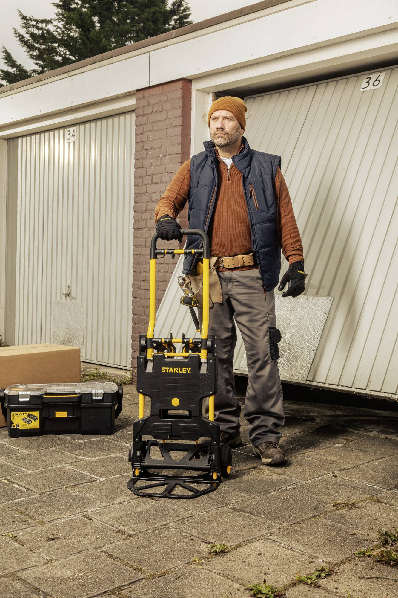 A tradesman is standing in front of a garage, holding a wheeled trolley with a toolbox. He is wearing a cap and work attire.