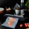 A vacuum sealer seals salmon pieces in a bag. In the background, a pot with herbs in water and tomatoes beside it.