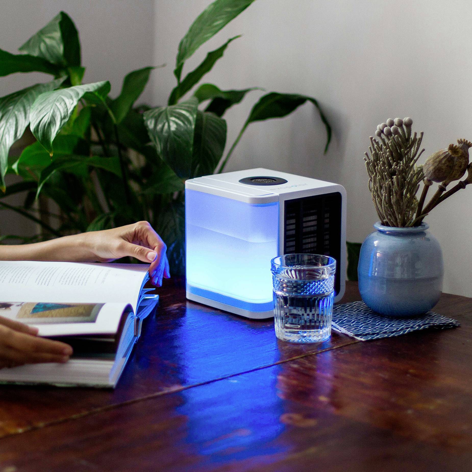 A person is reading a book at a table with a small, glowing air cooler, plants and a vase in the background.