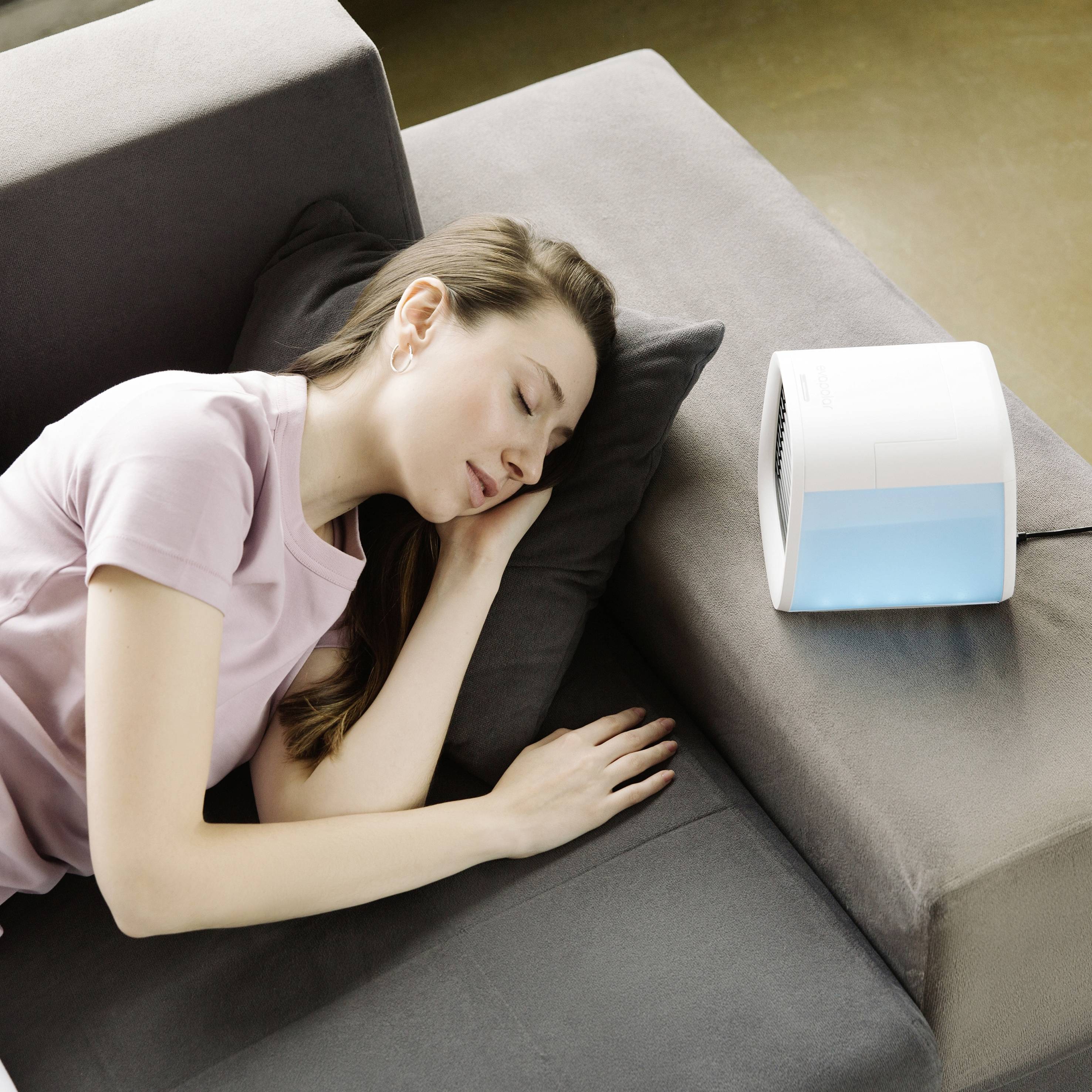 A woman is sleeping on a grey sofa next to a small, glowing air purifier, which creates a calming atmosphere.