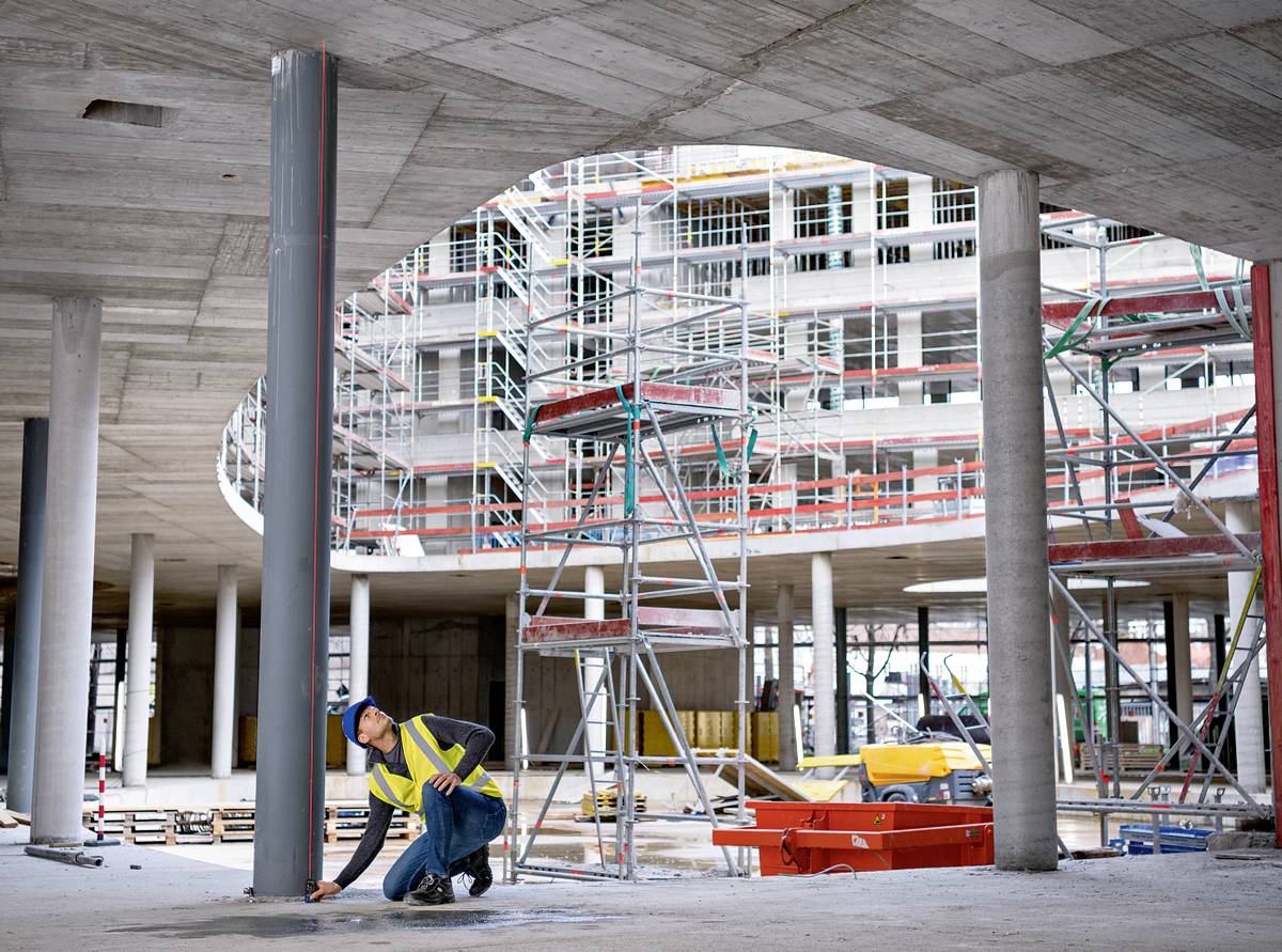 Construction worker kneels on building site, securing metal girder. In the background, a complex scaffolding system and an unfinished building.