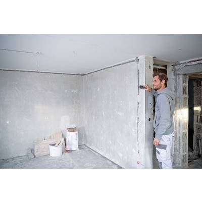 A man is measuring the height of a wall using a digital device in an unfinished room with bare walls and building materials.