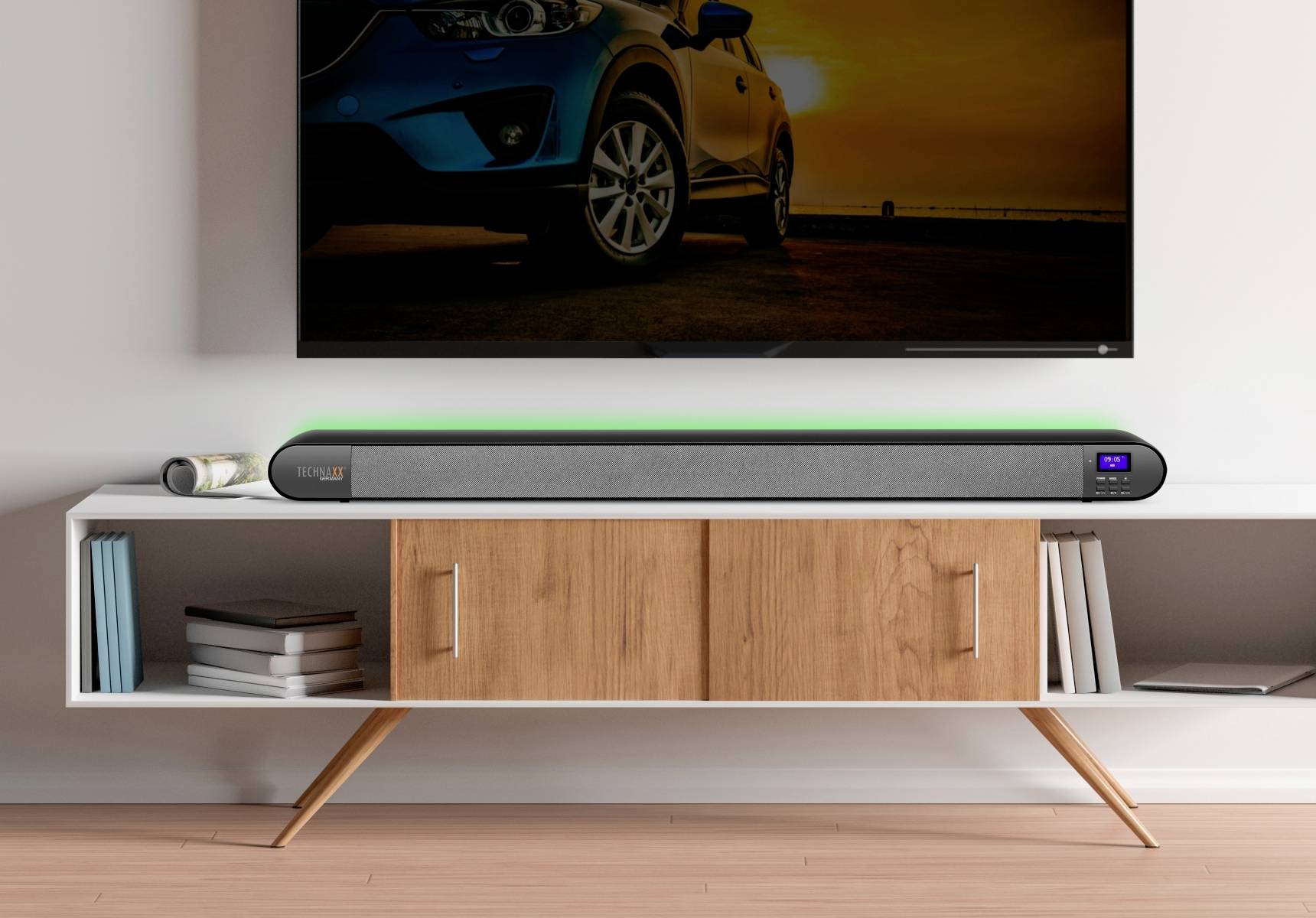 A grey soundbar on a wooden shelf beneath a wall-mounted television with a car in the twilight.